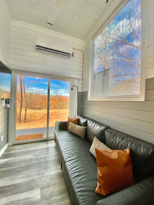 Cozy interior of a modern tiny home with a leather sofa and large windows overlooking a scenic outdoor view.